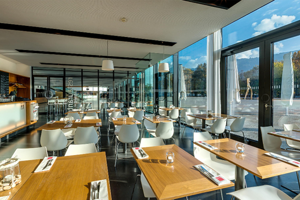 Holz-Tische mit weißen Stühlen, eine Ausgabetheke und eine lange Glasfront im Restaurant buehnedrei im Festspielhaus Bregenz