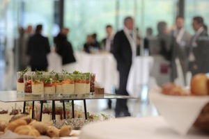Verschiedene Dessertgläschen und Gebäck, im Hintergrund stehen Kongressteilnehmer:innen an Stehtischen