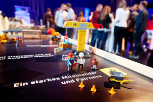 Playmobil Straßenbau mit Kran, im Hintergrund Menschen bei einem Empfang
