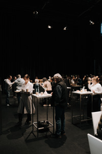 Menschen stehen an Hochtischen in einem dunklen Raum, Servicepersonal trägt Getränke und Speisen.