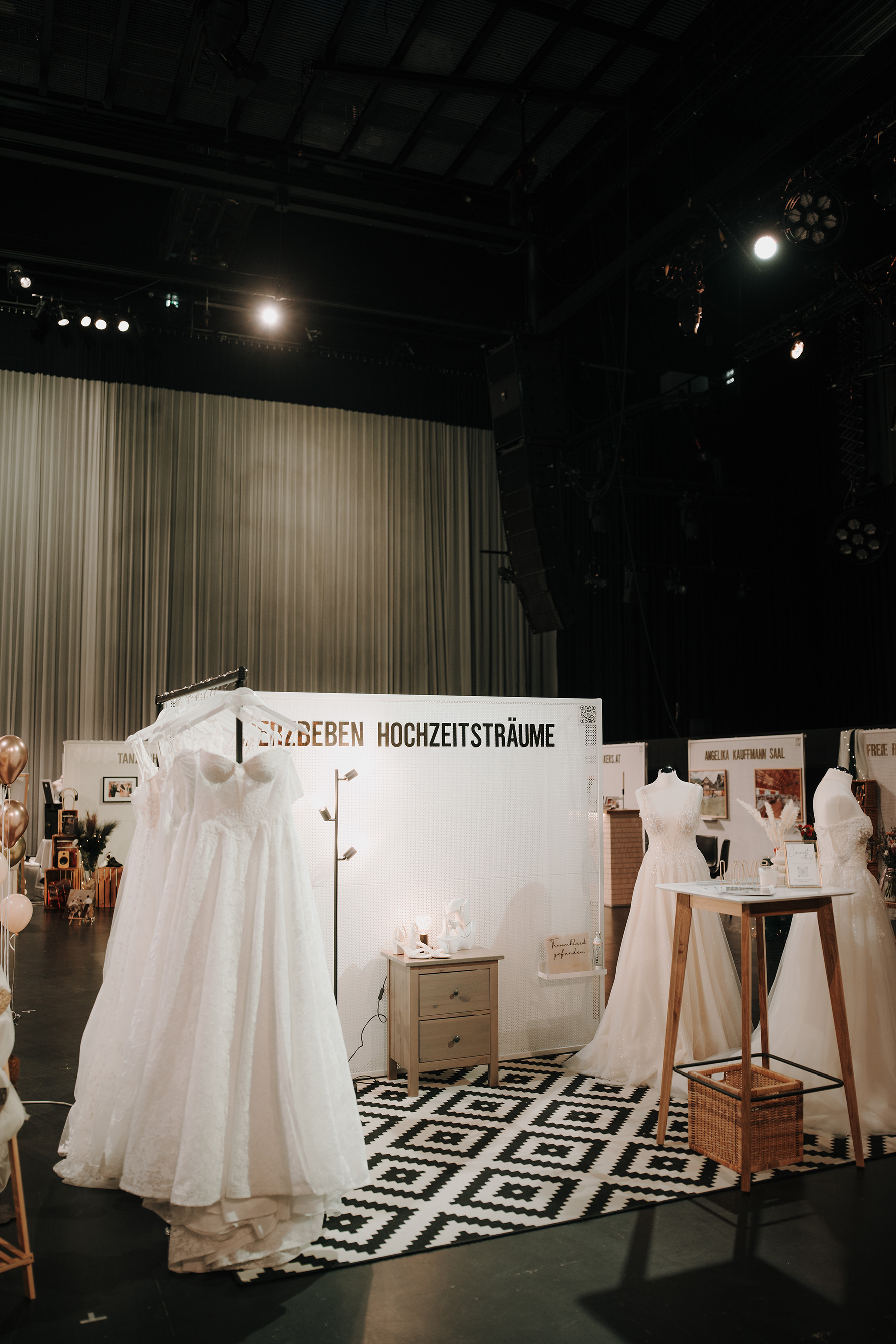 Mehrere Brautkleider hängen und stehen auf einer Ausstellungsfläche mit Teppich und Möbeln. © The CLICK. Wedding